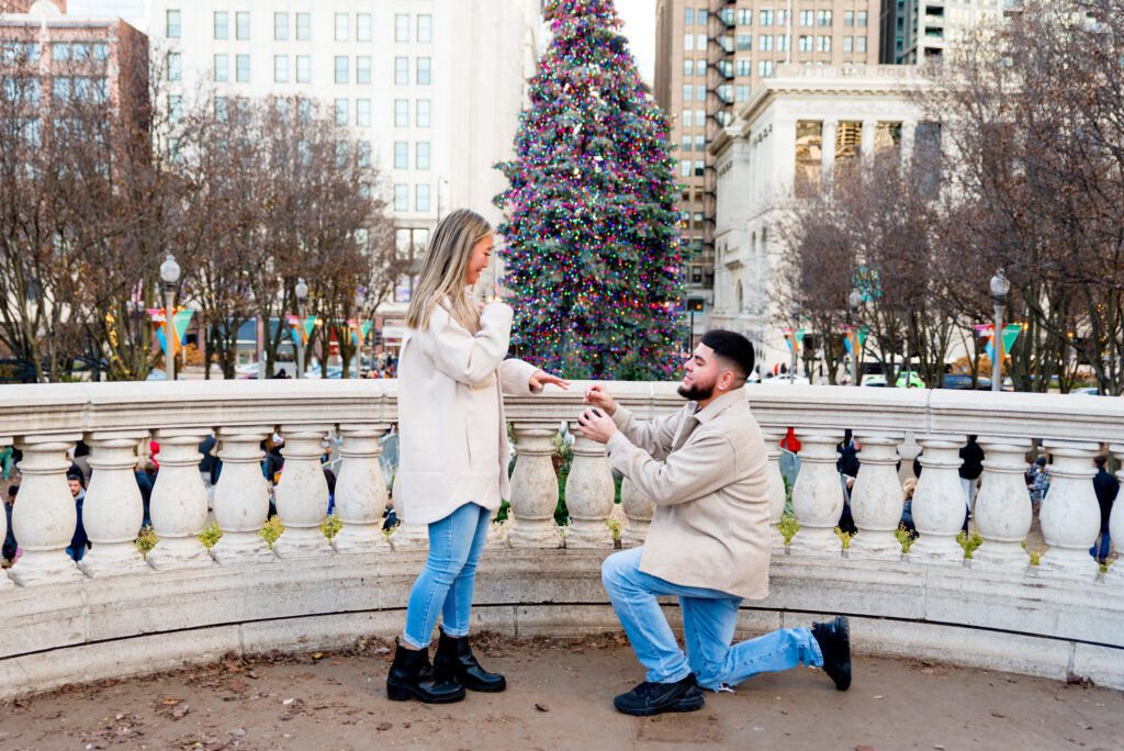 Man in tan jacket and blue jeans proposes to girlfriend in matching outfit in front of the colorful Christmas Tree in Grant Park in downtown Chicago IL. 