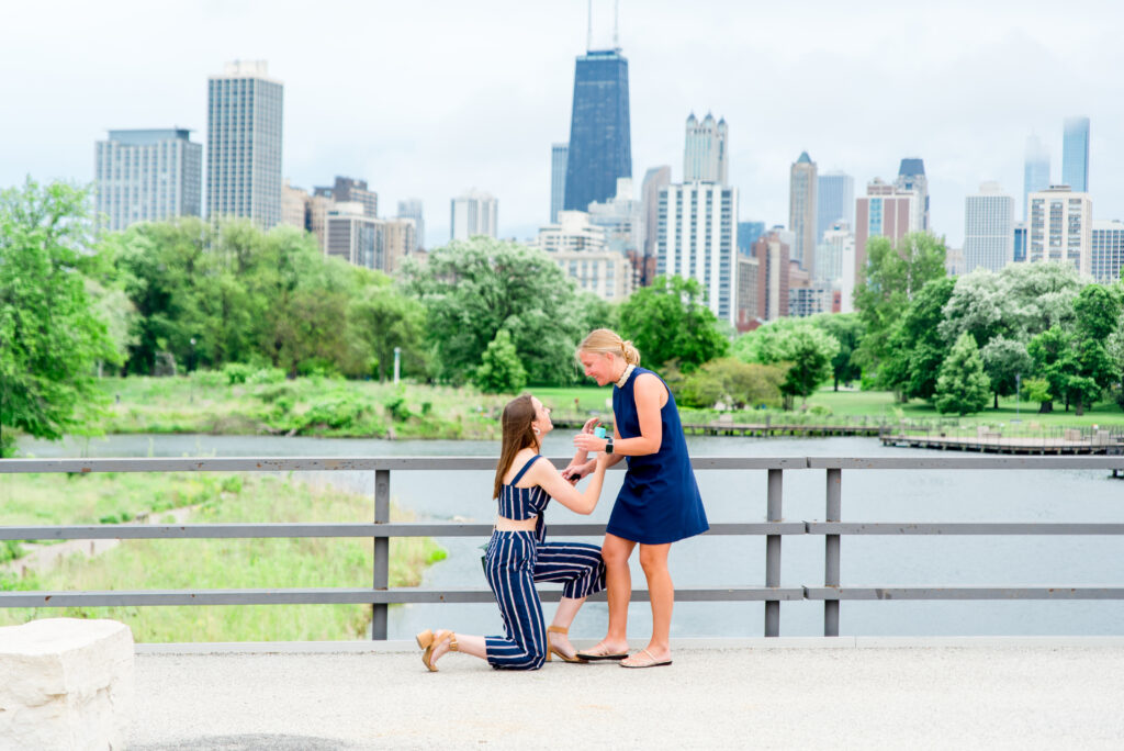 Lesbian couple in coordinating navy dress and jumpsuit are standing in front of the Chicago skyline view from Lincoln Park South pond, with one woman down on her knee proposing. 