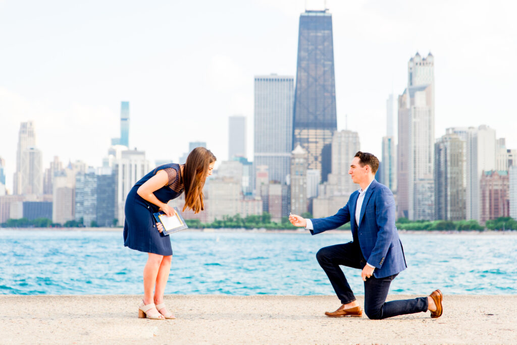 Woman is smiling at her fiance who just proposed at North Avenue Beach in Chicago during the summer. Both are in navy blue clothing and the Chicago skyline is behind them. 