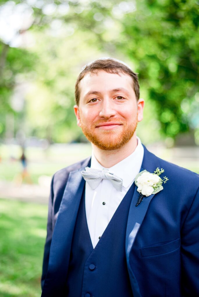 groom in navy suit and light gray bowtie with a white boutonniere smiles