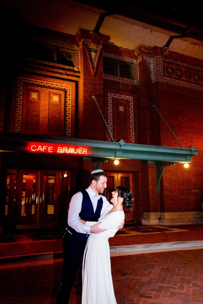 bride and groom hold each other and smile in front of Cafe Brauer at night with the red Cafe Brauer sign lit up 