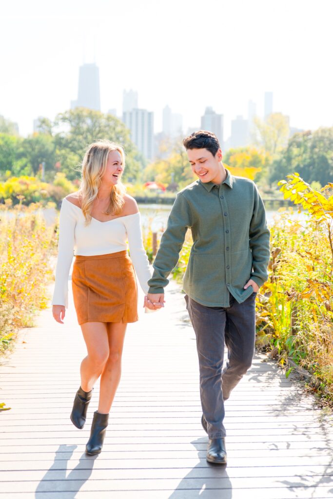 Man and woman in earth tones hold hands and walk on the path at Lincoln Park South Pond nature path in the fall.
