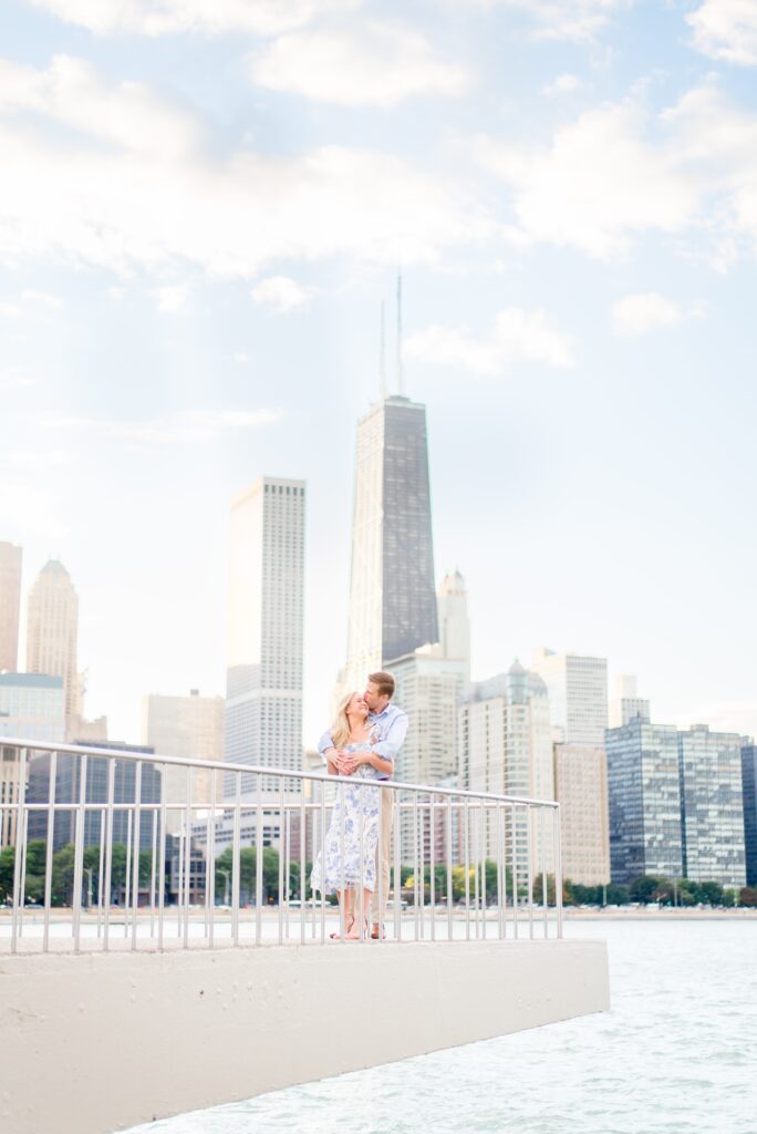 Man and woman in light blue hug each other in front of Chicago skyline at Milton Lee Olive Park for a summer proposal. They are standing on the edge of an overhang above the lake. 