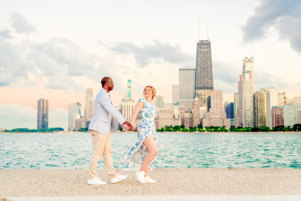 Woman in blue and white floral dress from Reformation walks along the pier at North Avenue Beach, holding hands and leading her fiance in a light blue jacket and tan pants. They walk in front of the Chicago skyline that's lighting up as the sunsets with clouds behind them. 