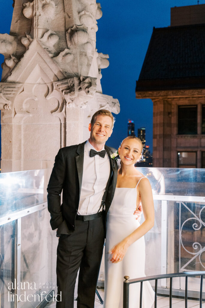 couples smiles on roof of University club of chicago at night with blue sky behind them 