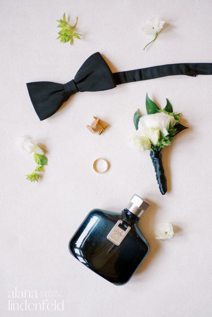 flat lay of black bow tie, black cologne, cufflinks and wedding band with a white boutonniere 