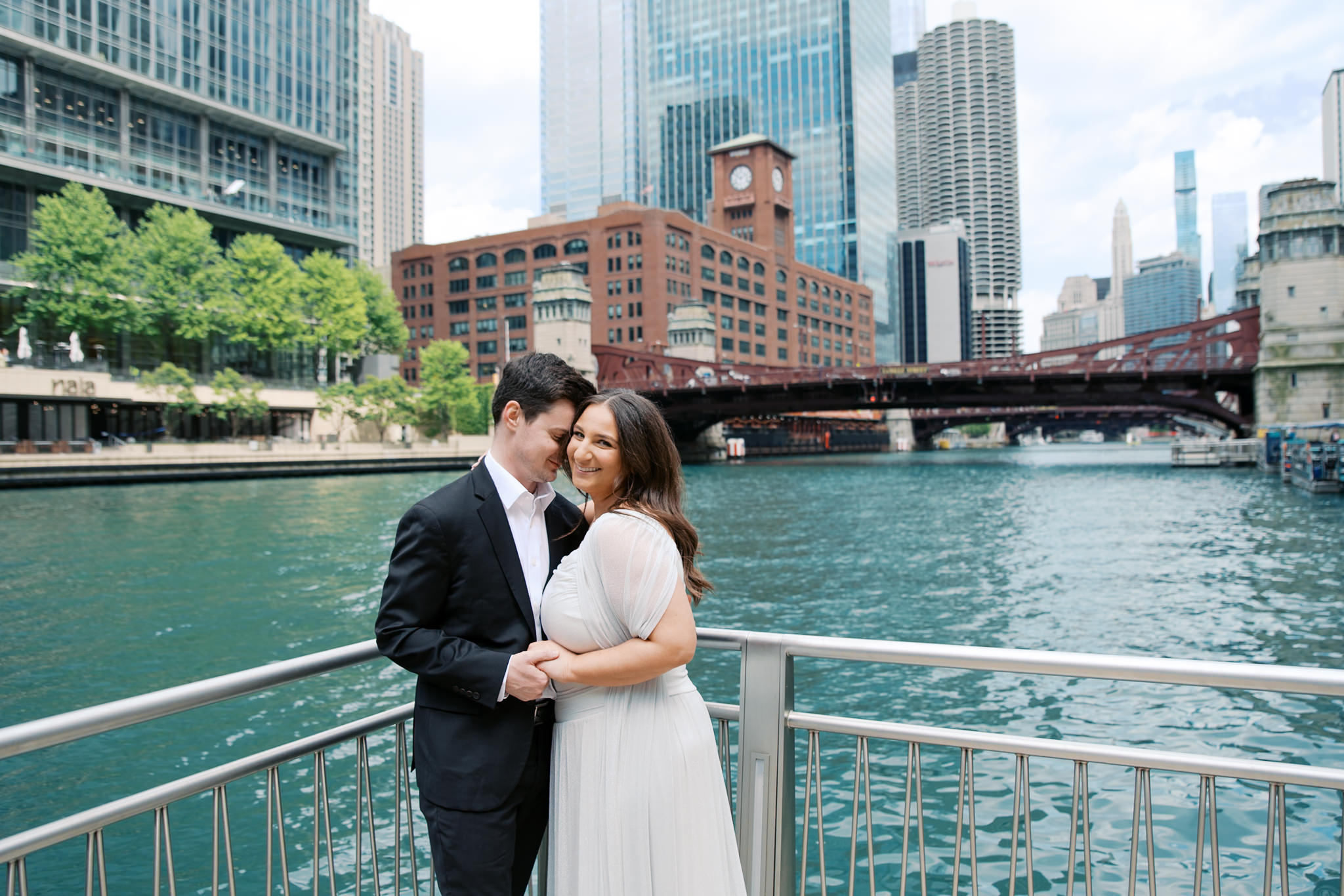 engagement picture in front of the Chicago Riverwalk on a summer day, with River Roast and LaSalle St. Bridge behind them