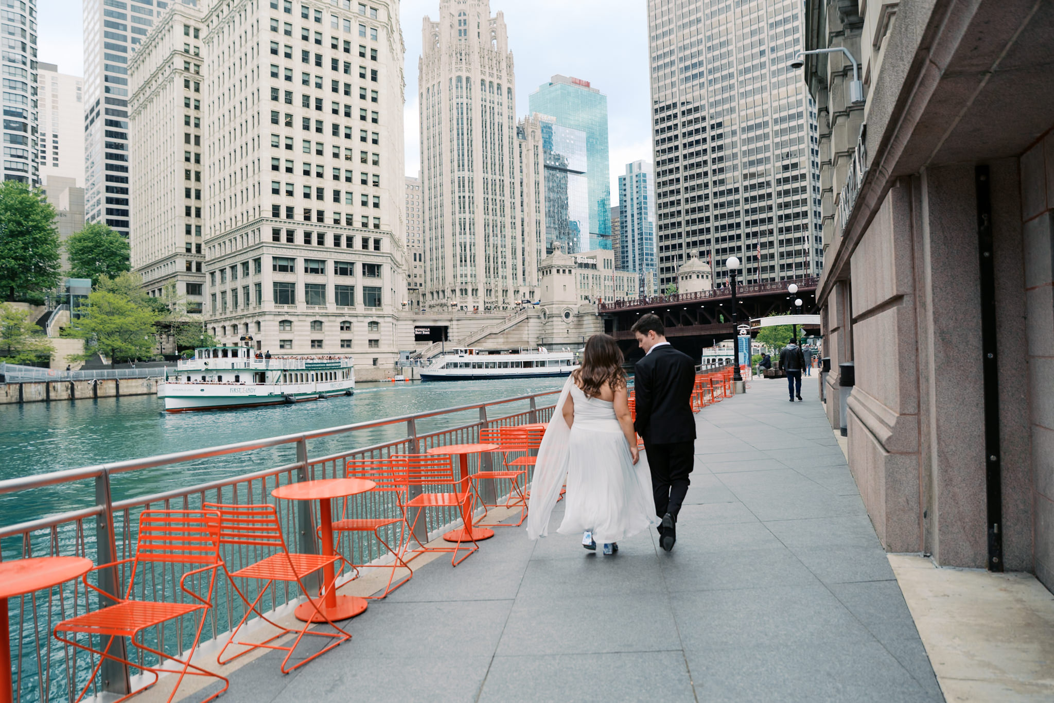 couple walking along chicago riverwalk on a summer day with tribune tower and wrigley building in the background